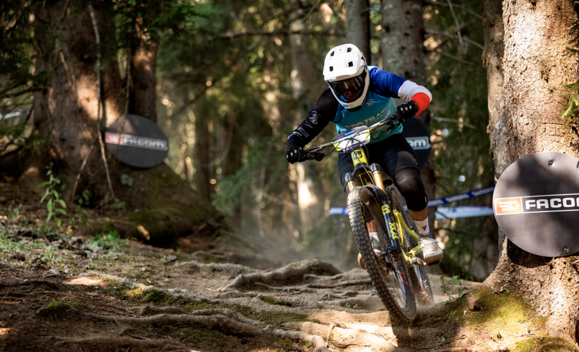 A mountain biker navigates a rugged, tree-lined trail filled with roots and dappled sunlight, showcasing an action shot that captures the intense focus and speed of the rider. Two sponsor signs are visible in the background, indicating a competitive race environment.