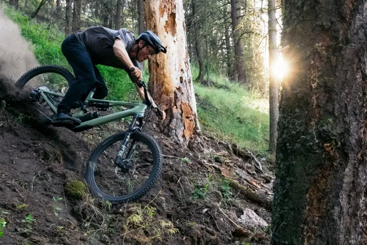 A mountain biker navigating a steep, dirt incline in a forested area, kicking up dust as the sun sets in the background, illuminating the trees.