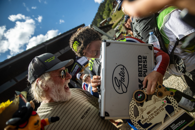 A joyful scene capturing a man with a white beard and sunglasses smiling broadly as he interacts with a young athlete wearing a bright racing outfit and a furry hat. The athlete is holding a large, silver case and a trophy with a detailed design, while a crowd and scenic mountain background can be seen behind them. The atmosphere conveys excitement and celebration.