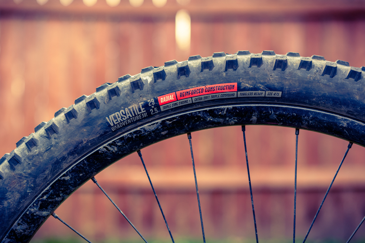 A close-up of a mountain bike tire showcasing its tread pattern and branding. The tire is labeled "Versatile Adventure" with additional specifications, including a size of 29 x 2.5, "Radial," "Reinforced Construction," and "Tubeless Ready." The background features a blurred wooden fence, emphasizing the tire's details.