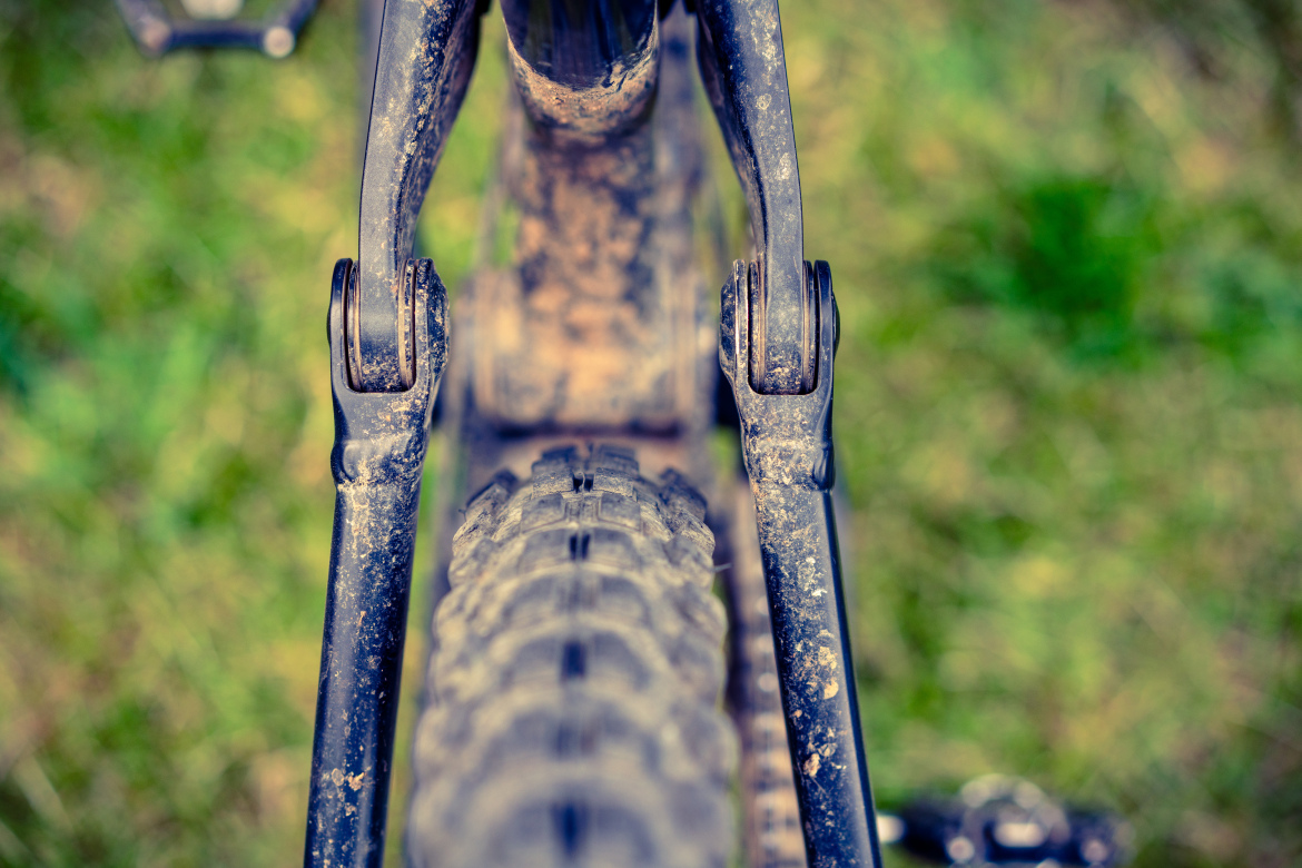 Close-up view of a mountain bike's rear suspension and tire, showcasing the muddy components against a blurred green grass background. The image emphasizes the intricate details of the bike's mechanics.