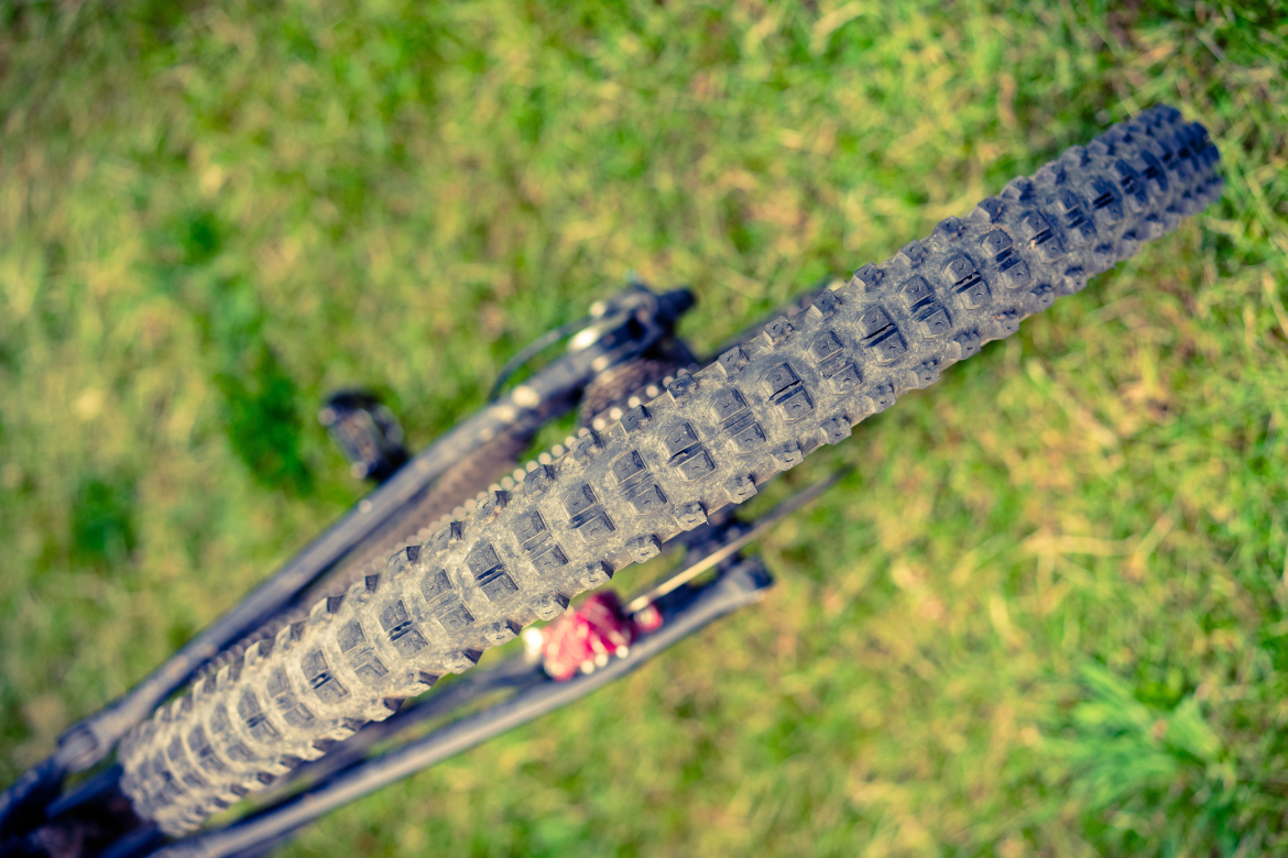 A close-up view of a mountain bike tire resting on green grass, showcasing the textured tread pattern. The bike's frame and components are partially visible in the background.