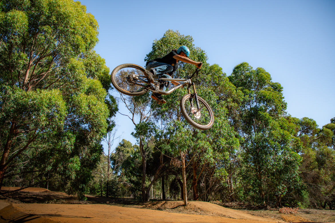 A person performing a mid-air jump on a mountain bike, surrounded by lush green trees and blue skies, showcasing a dynamic and adventurous outdoor scene.