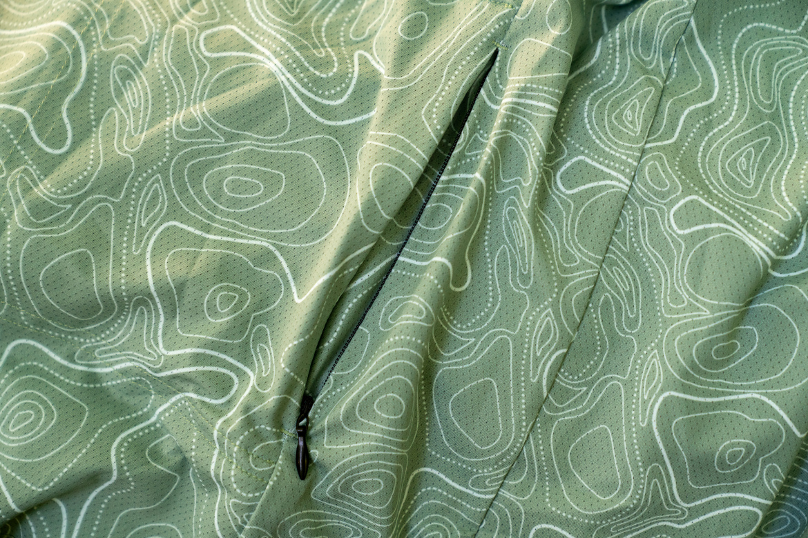 A close-up of a textured green fabric featuring a light-colored topographical pattern and a small zippered pocket.