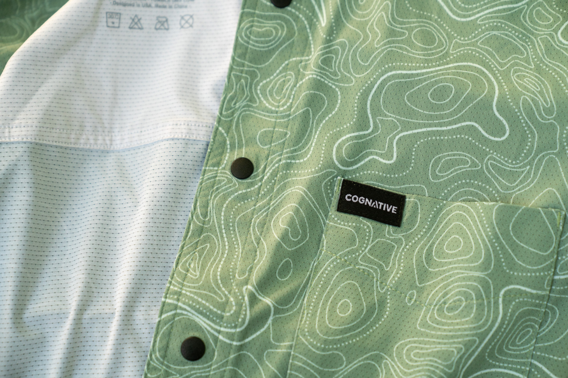 A close-up image of a green shirt featuring a topographic pattern and a black label that says "COGNITIVE." The shirt has a contrasting light blue fabric section, and visible buttons along the edge. The background is softly blurred to emphasize the details of the fabric and design.