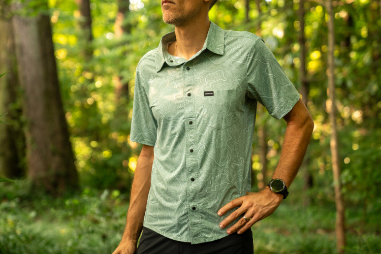 A person standing in a forest, wearing a light green short-sleeved shirt with a contour map design. The individual has their hand on their hip and is looking off to the side, with trees and greenery in the background.