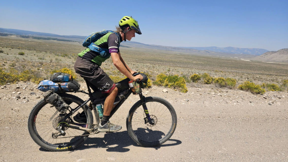 A person riding a mountain bike on a dirt path in an open landscape with sparse vegetation and distant hills under a clear blue sky. The rider is wearing a helmet and athletic gear, with a backpack secured on their back.