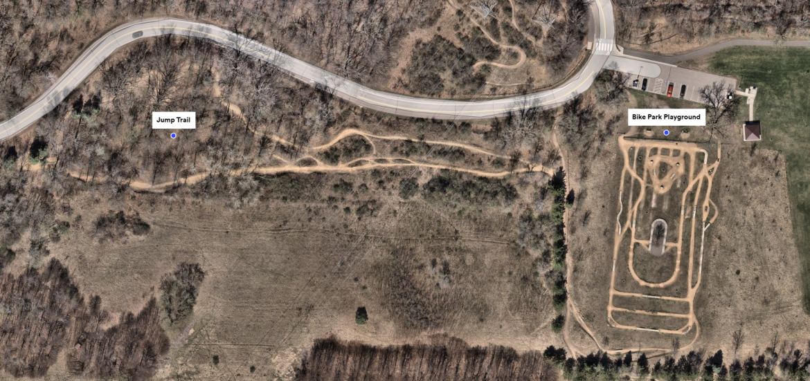 Aerial view of a park featuring a winding road, a designated bike park playground with a trail layout, and a nearby jump trail among a wooded area. The scene shows different dirt paths for biking and recreational space.