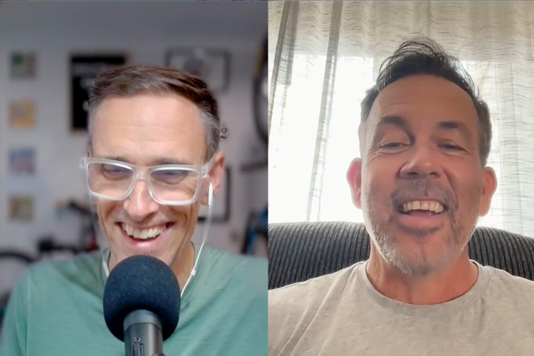 A split-screen video call featuring two smiling men. The man on the left, wearing glasses and a green shirt, speaks into a microphone. The man on the right, dressed in a light gray shirt, is seated in front of a window with sheer curtains, also smiling. The background includes blurred home office decor and a bicycle.