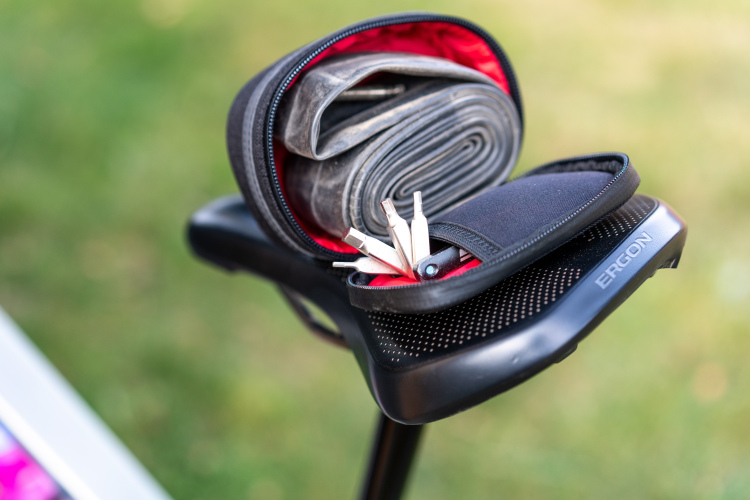 A bicycle seat with a small black storage bag attached, featuring a rolled tube and various tools partially visible inside. The bag is opened to reveal its contents against a blurred green grass background.