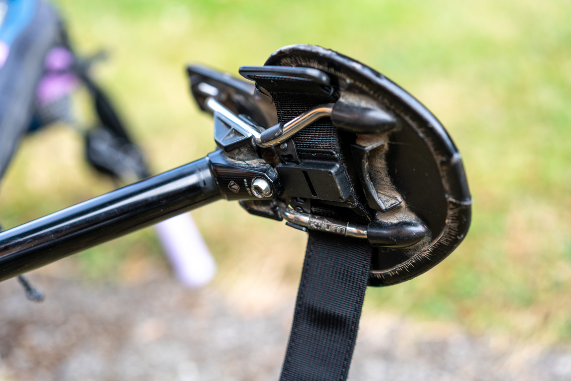 Close-up of the fastening mechanism on a trekking pole, featuring a sleek black handle, a securing clip, and a webbed strap. The background is a blurred green outdoor setting.