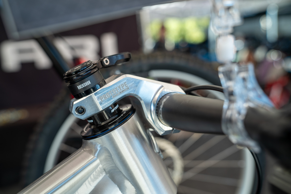 Close-up image of a bicycle's handlebar and stem, featuring a silver and blue prototype headset with a RockShox logo. The background includes blurred bicycle wheels and parts, indicating a cycling event or workshop setting.