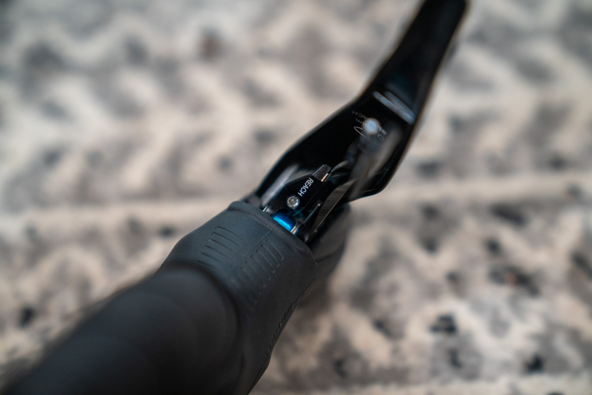 Close-up image of a bicycle brake lever with a textured black grip. The lever is partially angled, revealing internal components and a blue detail. The background features a subtle patterned surface.