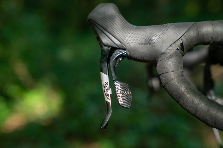 Close-up of a bicycle's handlebar featuring a SRAM gear shifter with a sleek design, wrapped in black tape for grip. The background shows a blurred green environment, suggesting an outdoor setting.