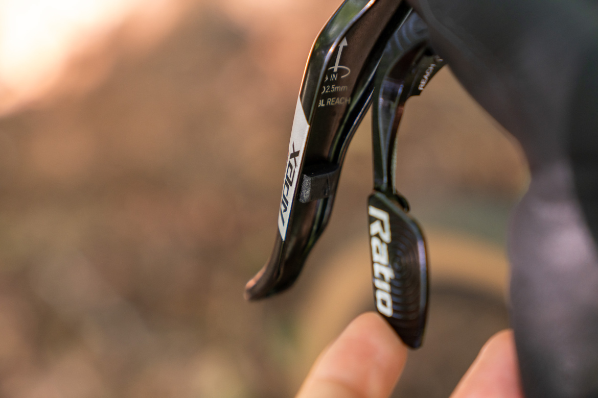A close-up image of a bicycle brake lever, featuring brand markings and adjustment indicators. A finger is seen gently touching the lever, which is set against a blurred natural background.