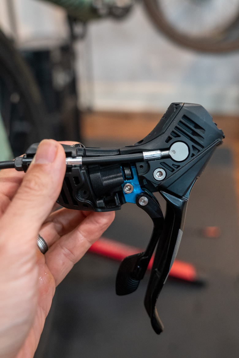 Close-up of a person's hand holding a bicycle gear shifter, showing intricate details and components. The shifter features a black plastic casing with visible mechanical parts, including a blue component and metal cables. The background includes a blurred view of bicycle parts and tools, suggesting a bicycle maintenance or repair setting.