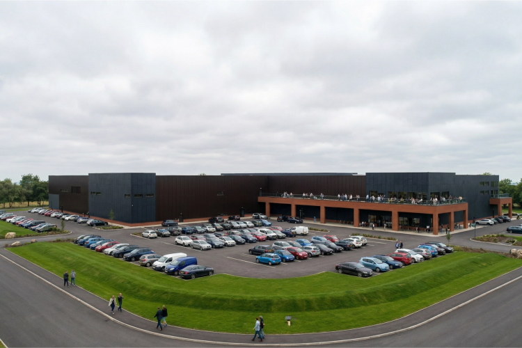 A modern commercial building with dark-colored exteriors, surrounded by a spacious parking lot filled with various vehicles. In the foreground, a landscaped area features manicured grass, and a few people are walking along the pathway. The sky is overcast, creating a muted atmosphere. The building has multiple entrances, and several people can be seen on an upper balcony.