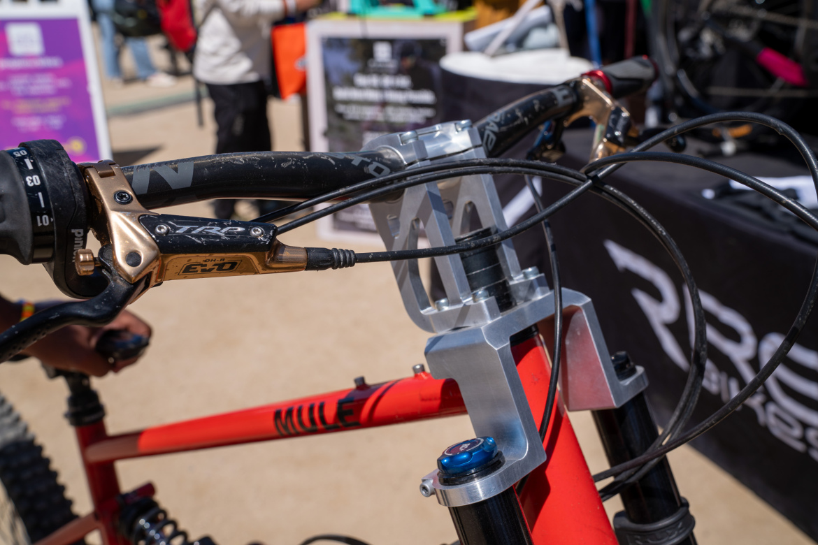 Close-up view of the handlebars and front suspension of a mountain bike, featuring black grips, a gold brake lever, and a silver component mounted on the bike frame. The background shows a blurred crowd and a black tent with a logo.