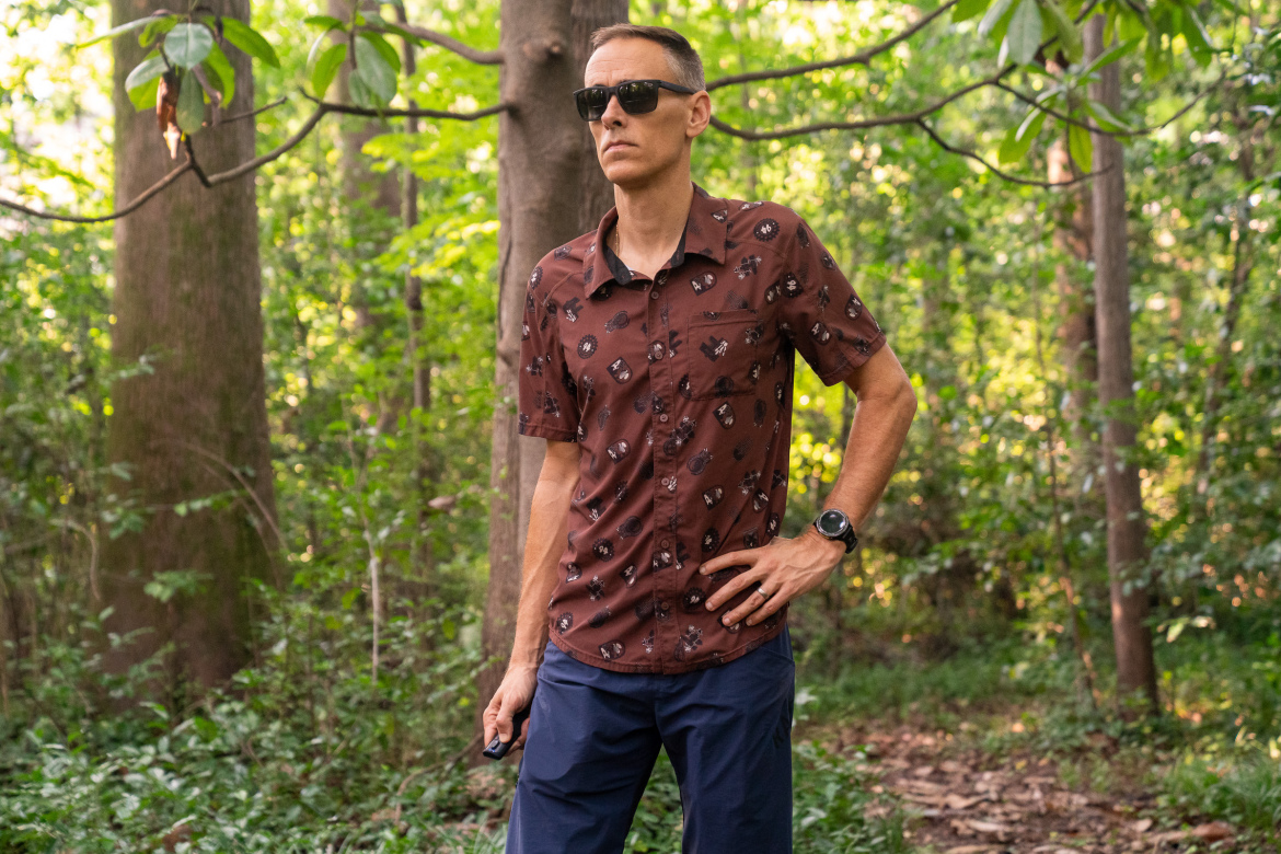 A person standing confidently in a forest, wearing sunglasses and a brown patterned shirt complemented by navy blue shorts. They hold a small object in their left hand and have their right hand on their hip, surrounded by lush green trees and underbrush.