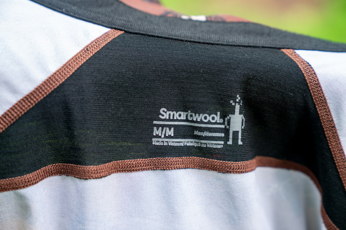 Close-up view of the back of a shirt featuring the Smartwool logo, along with size details and care information. The shirt has a color-block design with black, white, and brown sections. The label indicates it is made in Vietnam.