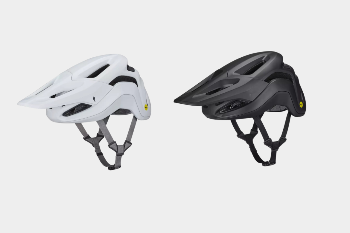 Two bicycle helmets are displayed side by side: one is white and the other is black. Both helmets feature a sleek, aerodynamic design with ventilation openings and adjustable straps. The white helmet has a prominent visor while the black helmet has a slightly different shape, showcasing modern safety design.