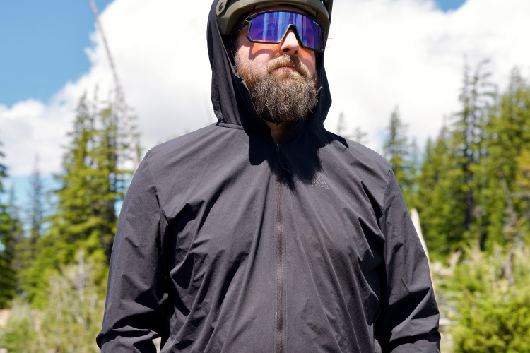 A person wearing a black zip-up jacket with a hood, paired with a helmet and sunglasses, stands in an outdoor setting with lush green trees and a partly cloudy sky in the background.