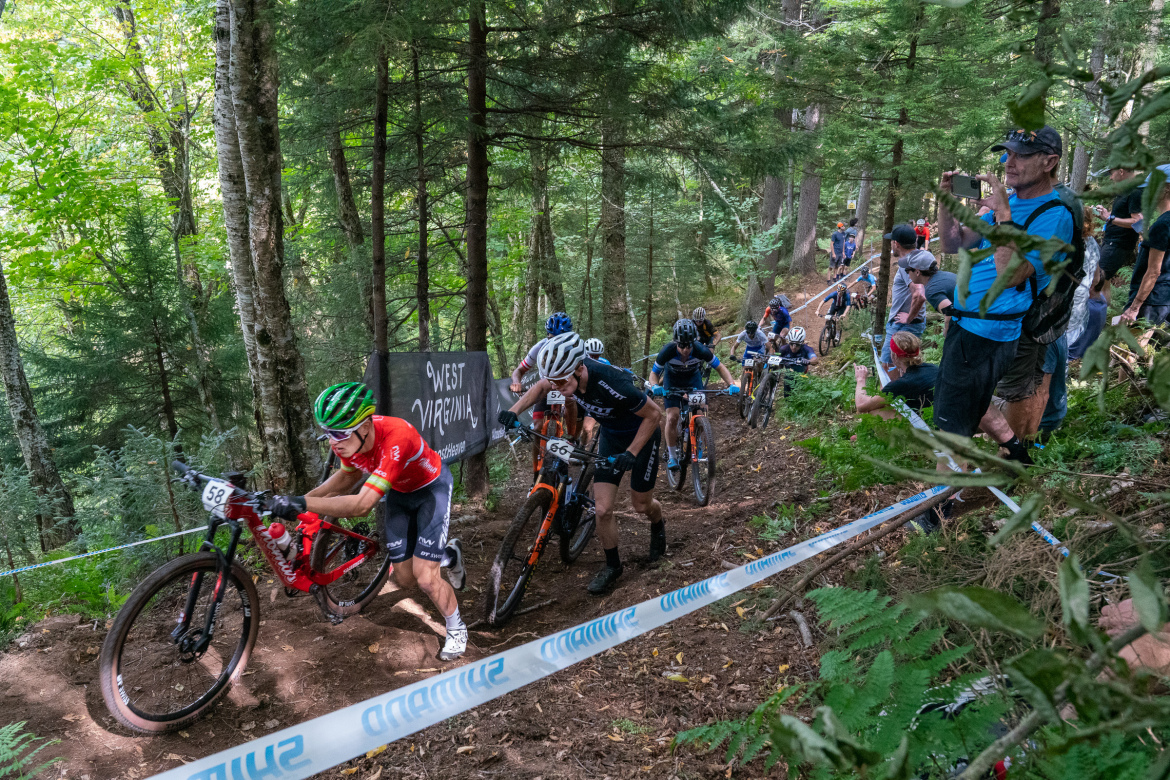 A group of mountain bikers navigating a steep and winding trail in a forested area. One cyclist in a red jersey is prominently in the foreground, while others follow closely behind. Spectators are visible along the course, cheering them on. A banner reading "West Virginia" is displayed in the background, indicating the location of the event. The lush greenery and trees provide a vibrant backdrop to the intense competition.