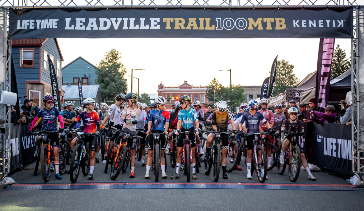 A group of mountain bikers lined up at the start line of the Leadville Trail 100 MTB race, with a large banner reading "LIFE TIME LEADVILLE TRAIL 100 MTB" overhead. The riders, wearing various team jerseys and helmets, are positioned on their bikes, ready for the race to begin amidst a backdrop of early morning light and spectators in the background.