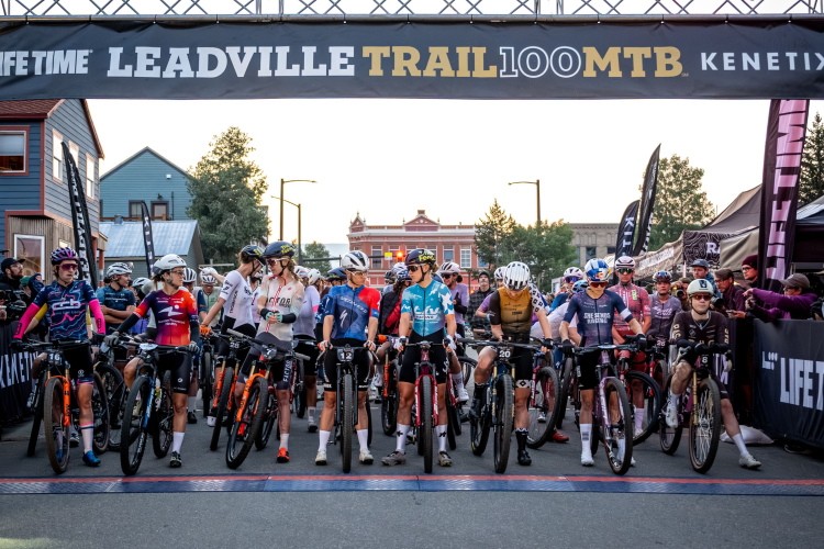 A group of mountain bikers lined up at the start line of the Leadville Trail 100 MTB race, with a large banner reading "LIFE TIME LEADVILLE TRAIL 100 MTB" overhead. The riders, wearing various team jerseys and helmets, are positioned on their bikes, ready for the race to begin amidst a backdrop of early morning light and spectators in the background.