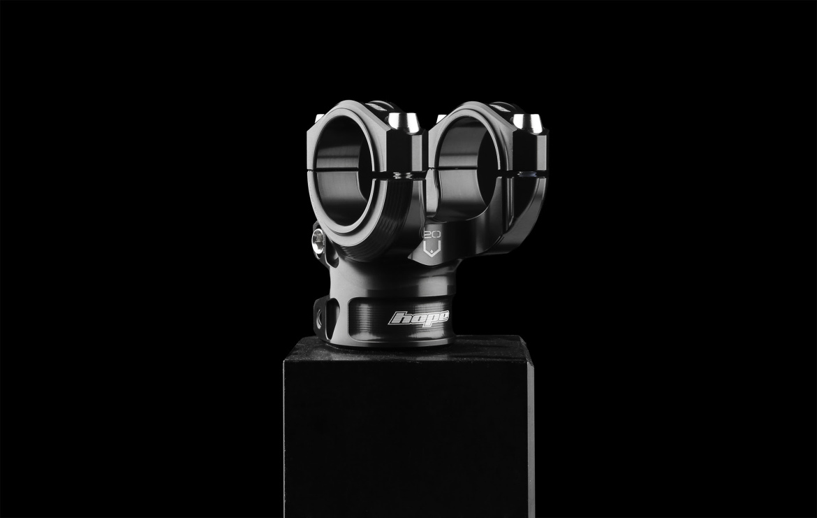 A sleek, black bicycle stem with a dual clamp design displayed on a pedestal against a dark background. The stem features a polished finish and the brand name "Hope" visible on its side.