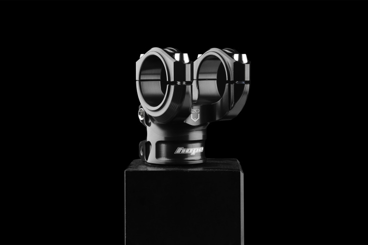 A sleek, black bicycle stem with a dual clamp design displayed on a pedestal against a dark background. The stem features a polished finish and the brand name "Hope" visible on its side.