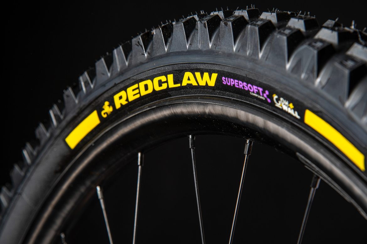 Close-up image of a mountain bike tire labeled "REDCLAW SUPERSOFT," featuring a textured tread pattern and a black background. The tire has distinctive yellow and purple branding, highlighting its design for enhanced grip and performance.
