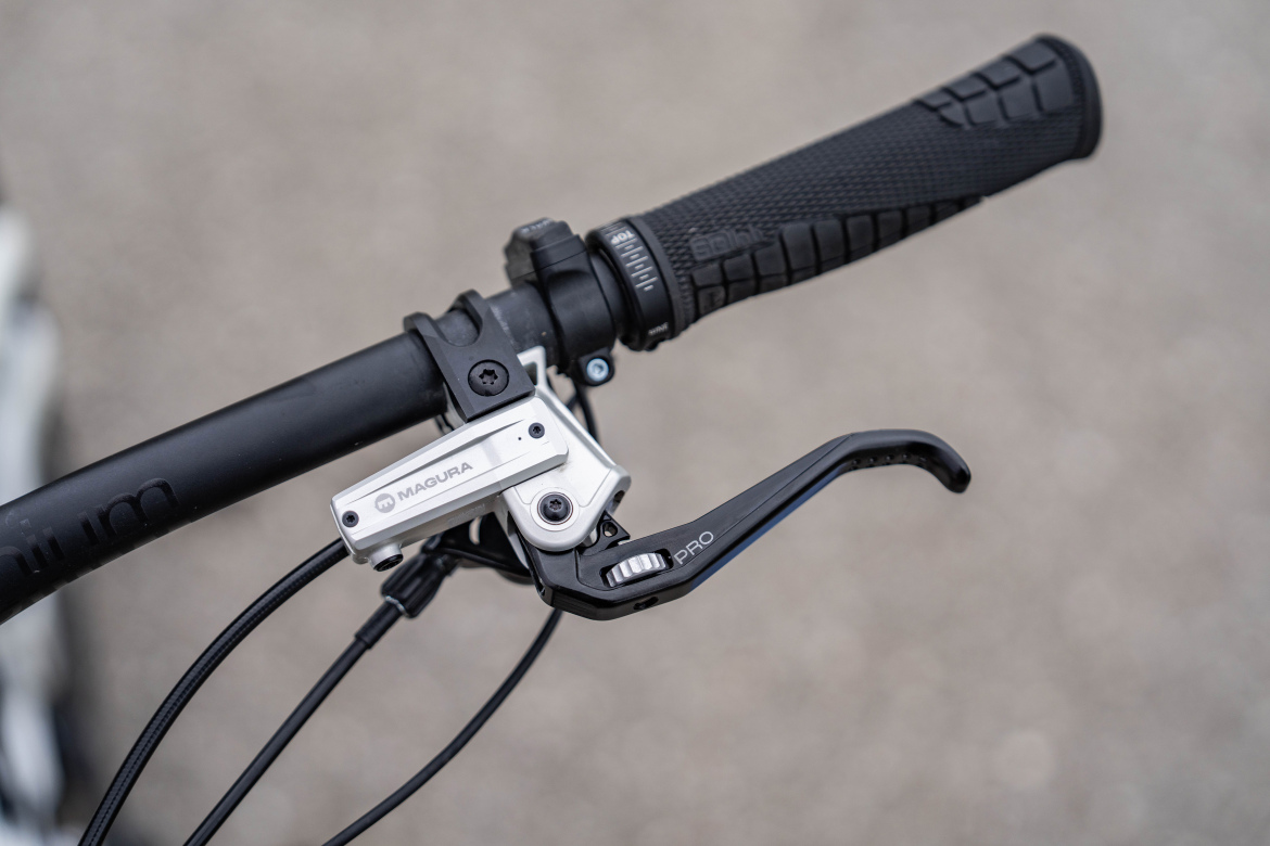 Close-up of a bicycle handlebar showing a brake lever and a grip. The brake lever is silver and black, with a logo visible, mounted on a black handlebar with textured grips. The background is a blurred, neutral surface.