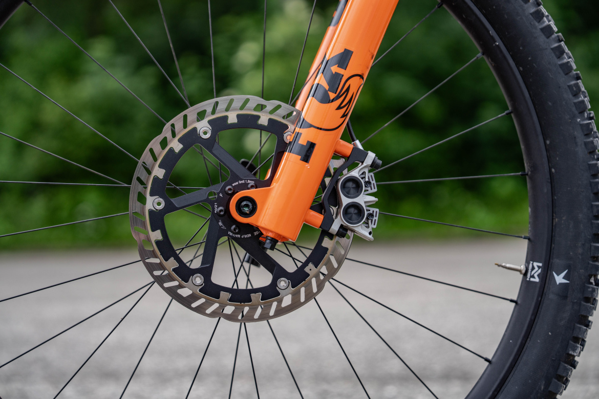 Close-up view of a mountain bike's front wheel, featuring a vibrant orange fork and a disc brake system. The black spokes and tire are visible, with a blurred green background, indicating an outdoor setting.