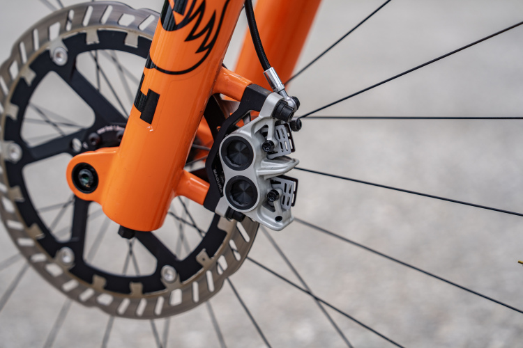 Close-up image of a bicycle's front wheel hub, showcasing an orange fork, a silver disc brake caliper, and a round perforated disc brake. The background is blurred, focusing on the bike's components.
