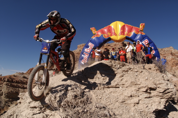 A mountain biker in a red and black jersey jumps off a rocky terrain, with a Red Bull inflatable arch in the background and a group of spectators watching nearby. The scene is set against a clear blue sky.
