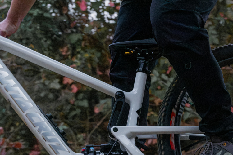 A close-up view of a cyclist's leg positioned on the frame of a white mountain bike, with a focus on the bike's rear suspension and components. The background features foliage with autumn colors. The cyclist is wearing dark pants and shoes, highlighting active outdoor gear.