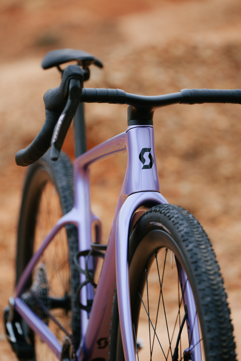 A close-up view of a sleek purple bicycle frame, showcasing the handlebars and front fork. The bike features textured tires suitable for gravel terrain, with a prominently displayed logo on the frame. The background consists of a blurred earthy landscape, highlighting the bike's design and color.