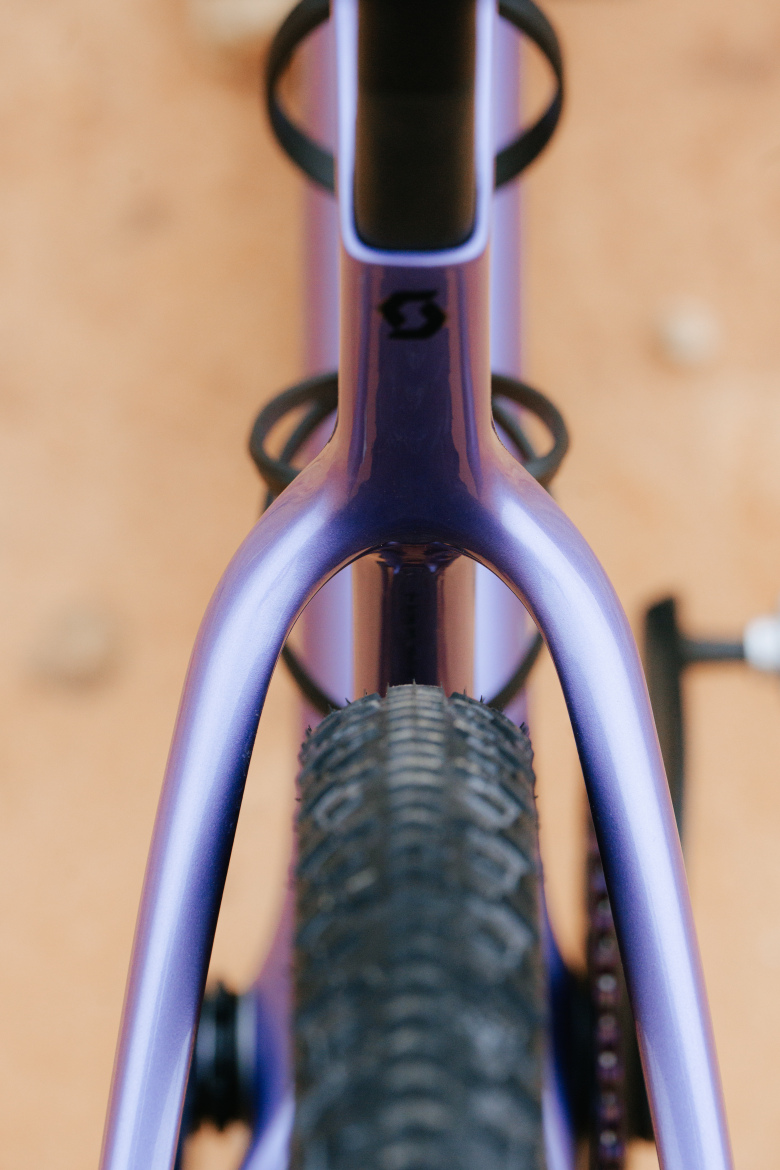 Close-up view of a purple bicycle frame, focusing on the top tube, seat post, and rear tire. The image highlights the sleek design and texture of the frame, with a blurred background of a dirt surface.