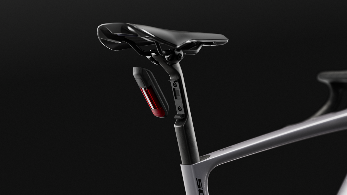 Close-up image of a bicycle saddle and seat post, featuring a sleek design with a red rear light attachment for enhanced visibility. The background is dark, emphasizing the modern contours and materials of the saddle and post.