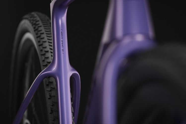 Close-up of a purple bicycle frame, showcasing the rear triangle and tire. The frame has a modern design with a smooth finish, and features text that reads &ldquo;addict gravel 10&rdquo; on the side. The background is black, enhancing the vibrant color of the bike.