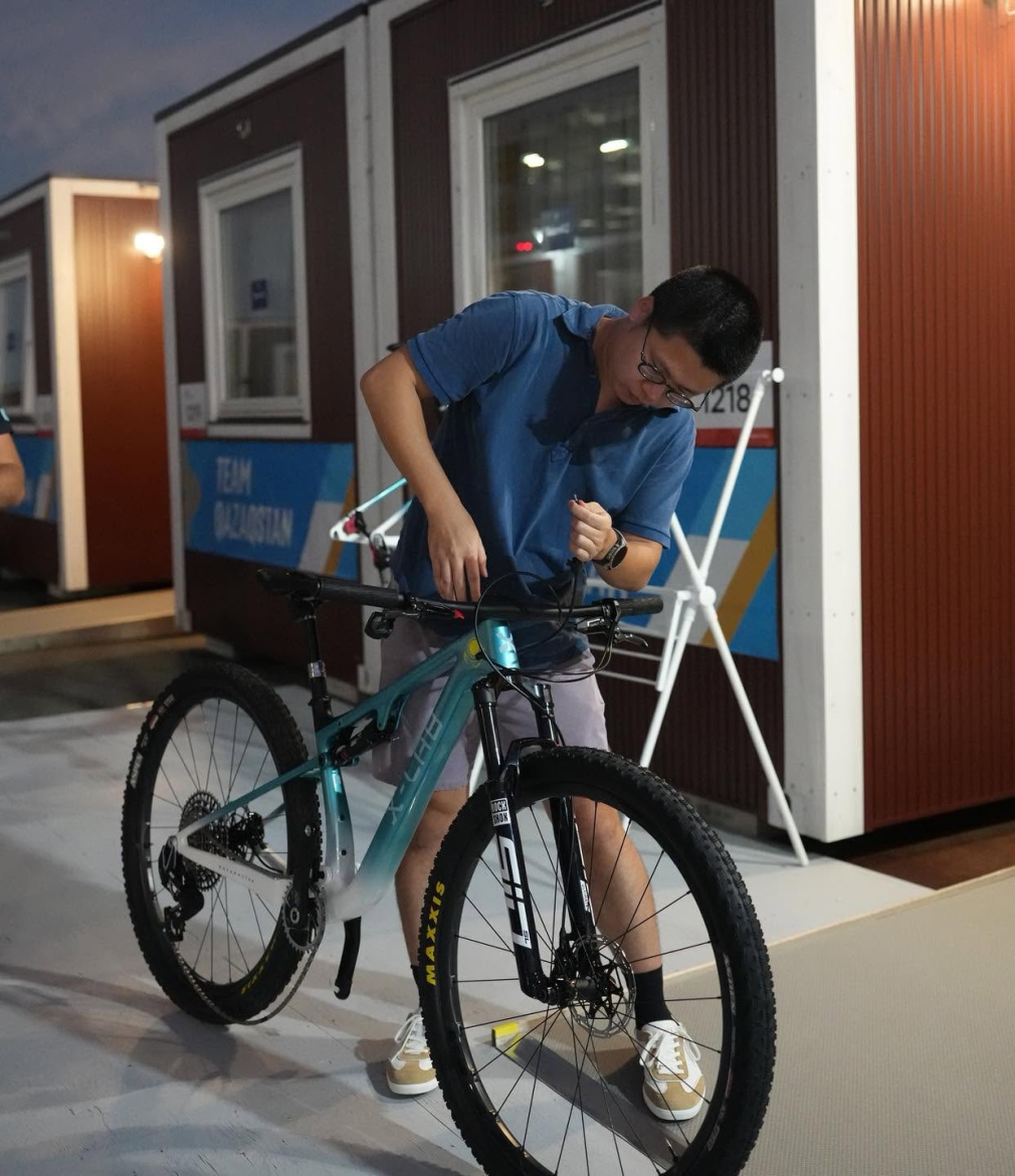 A young man wearing a blue polo shirt is adjusting the handlebars of a mountain bike in an outdoor setting at night. In the background, there are shipping container-style structures with colorful team signage. The area is well-lit, highlighting the bike and the individual engaged in maintenance.