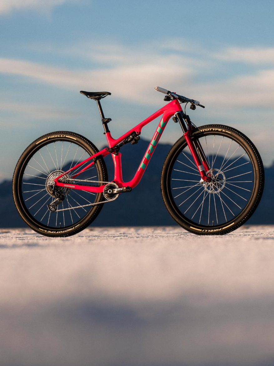A vibrant pink mountain bike with turquoise accents is positioned against a scenic backdrop featuring a clear sky and distant mountains. The bike features modern components, including disc brakes and a lightweight frame, set on a smooth surface.