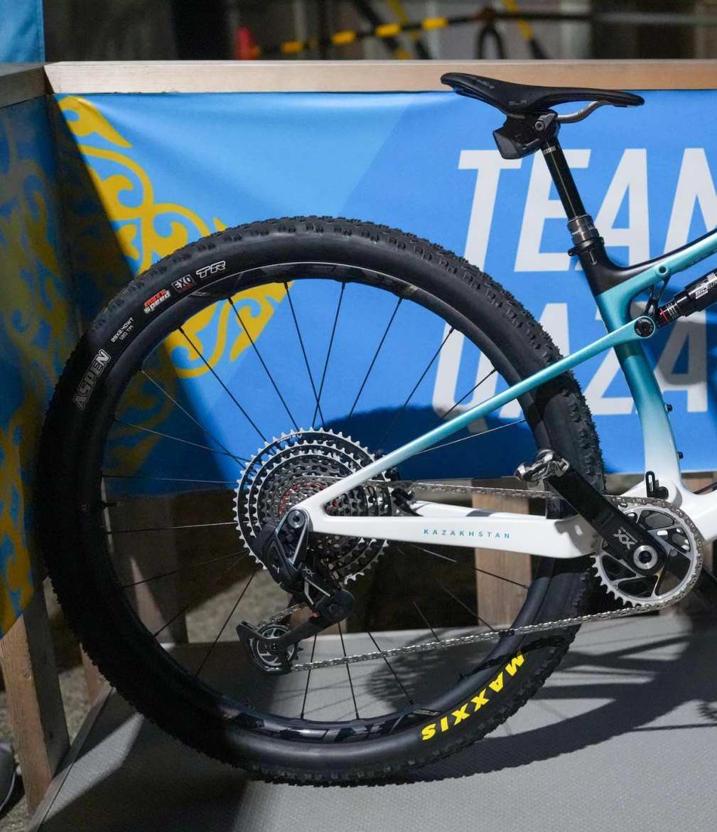 A close-up view of the rear wheel and drivetrain of a mountain bike, featuring a sleek frame painted in blue and white, a prominent "Kazakhstan" label, and black Maxxis tires. The background includes a blue banner with yellow patterns.