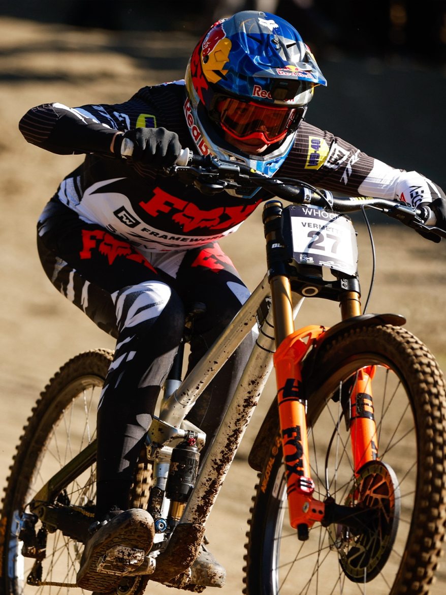 A competitive mountain biker wearing a blue helmet, goggles, and a full-body racing outfit speeds down a dirt trail, showcasing dynamic motion and focus. The bike features a distinct frame with mud splatter, emphasizing the rugged terrain.