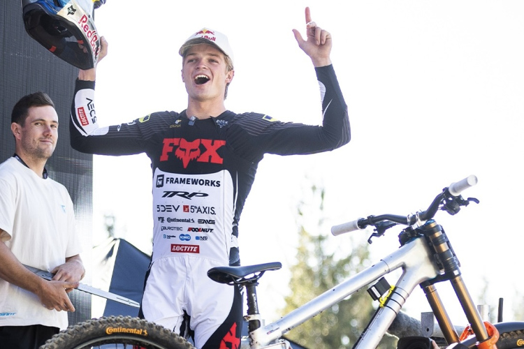 A mountain biker celebrating on a podium, wearing a black and white racing outfit with logos, including "FOX" and "Red Bull." He holds a helmet and raises one hand in triumph. In the background, another person stands nearby, and a mountain bike is positioned next to him. The setting is outdoors under bright sunlight.