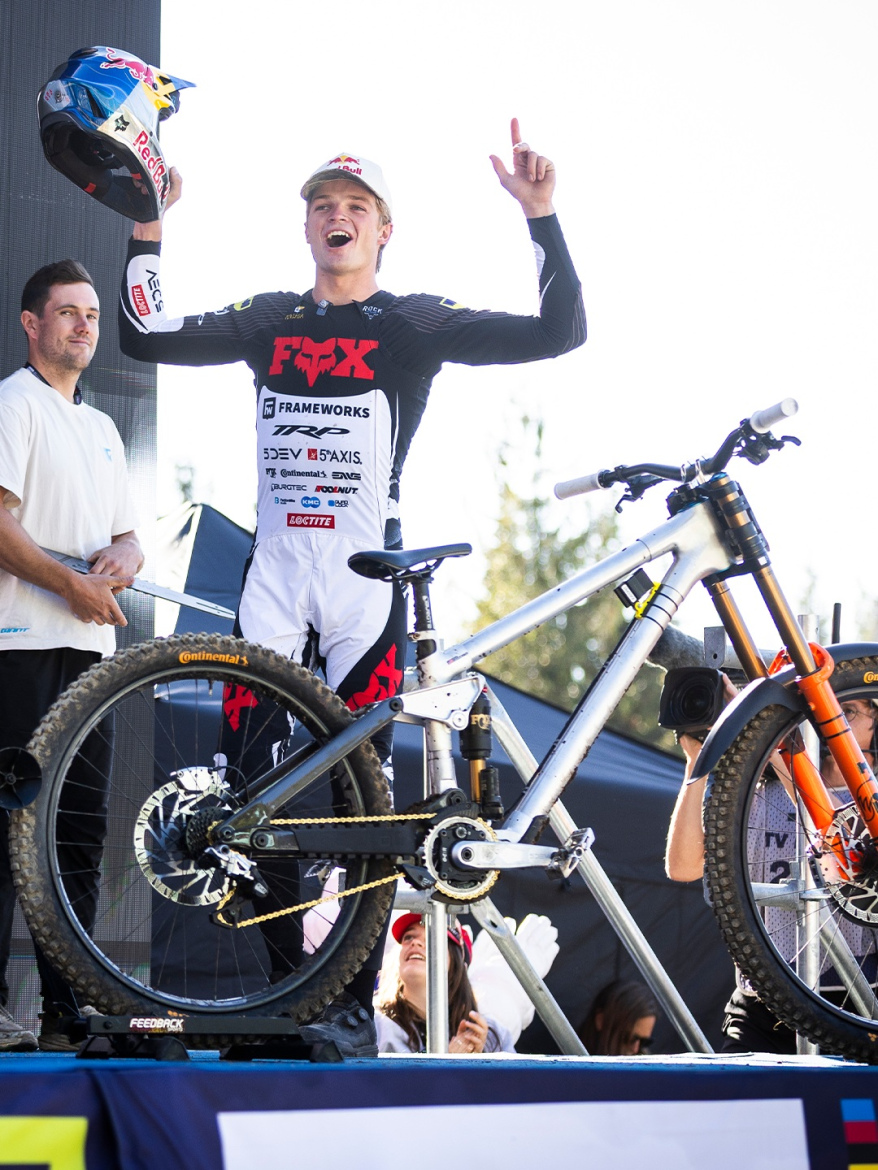A cyclist celebrating a victory on a podium, holding a helmet in one hand and raising a finger in triumph while wearing a racing outfit with branding. In the foreground, a mountain bike is prominently displayed. A supporter stands nearby, and the background features cheering spectators and event signage.