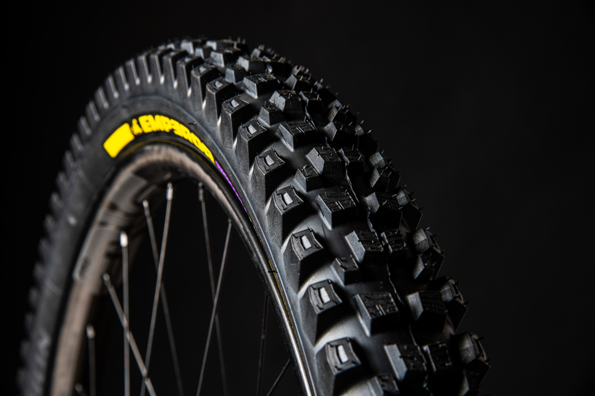 A close-up view of a mountain bike tire featuring a rugged tread pattern, designed for optimal traction on various terrains. The tire is partially turned to showcase its profile, with a black background highlighting the details of the tread and sidewall markings.