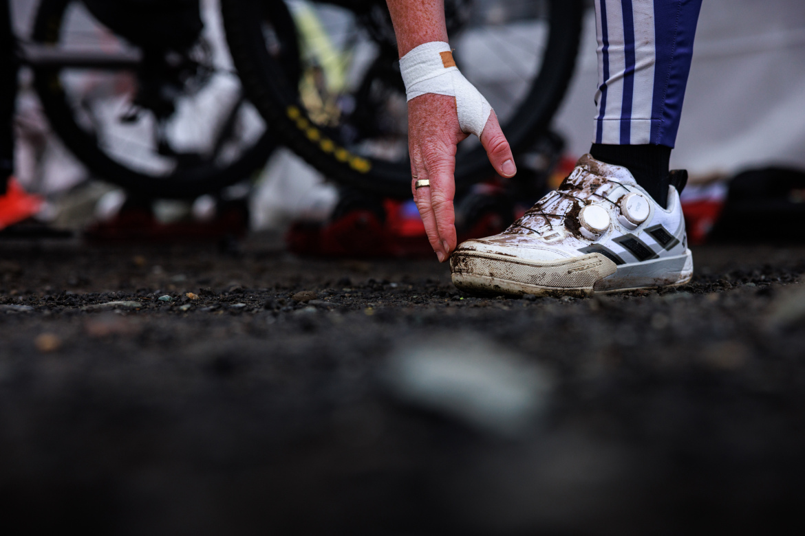 A close-up of a hand reaching towards a muddy white athletic shoe, set against a background of gravel and blurred bicycle wheels. The hand is bandaged, and the shoe features a dial adjustment system.
