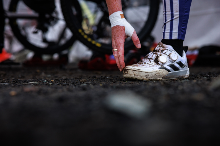 A close-up of a hand reaching towards a muddy white athletic shoe, set against a background of gravel and blurred bicycle wheels. The hand is bandaged, and the shoe features a dial adjustment system.